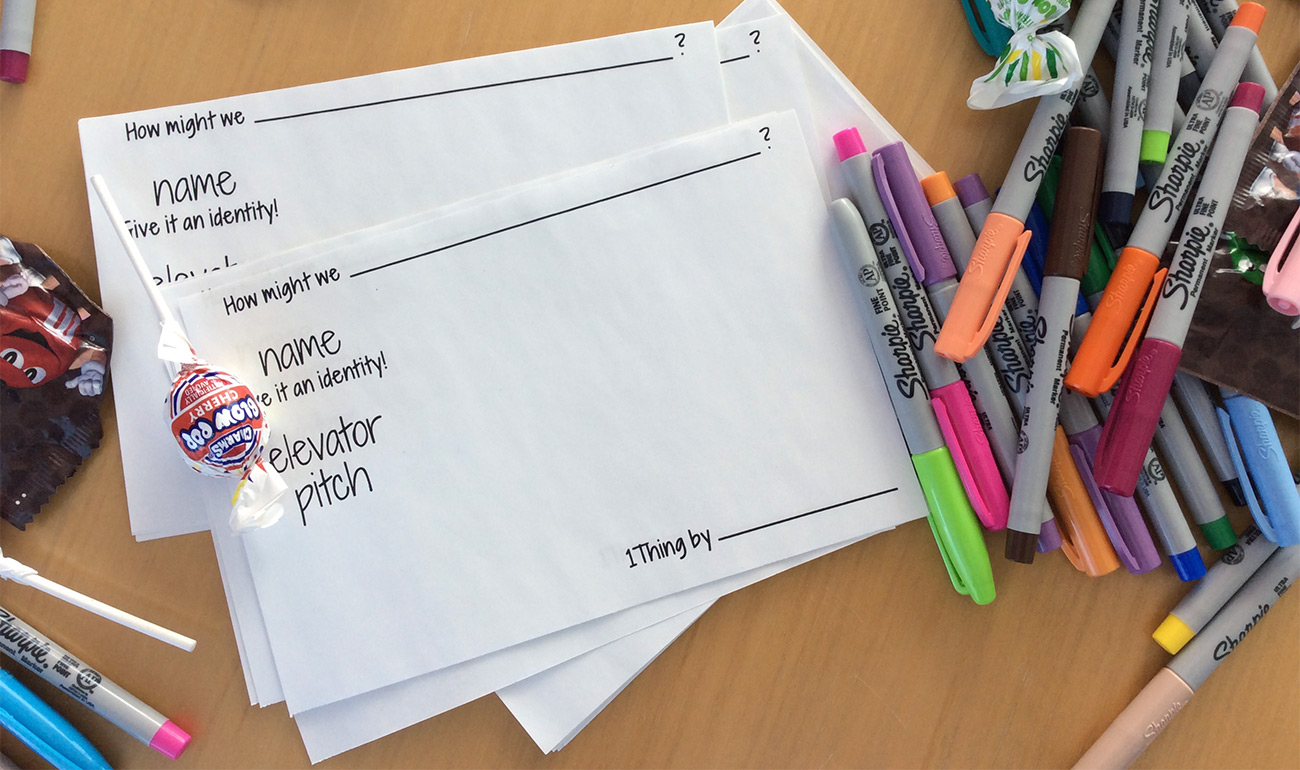 White cards with "how might we" statements on a wood table with colorful felt-tip markers and candy