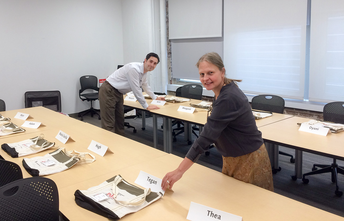 A woman and a man stand by desks with name tags, arranging papers and other classroom workshop materials
