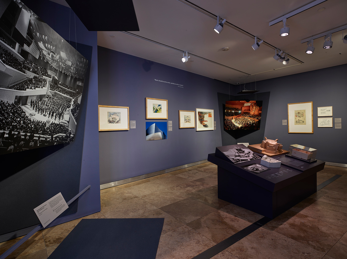 Interior of a gallery with dark blue walls, architectural models, and small watercolors and renderings in frames