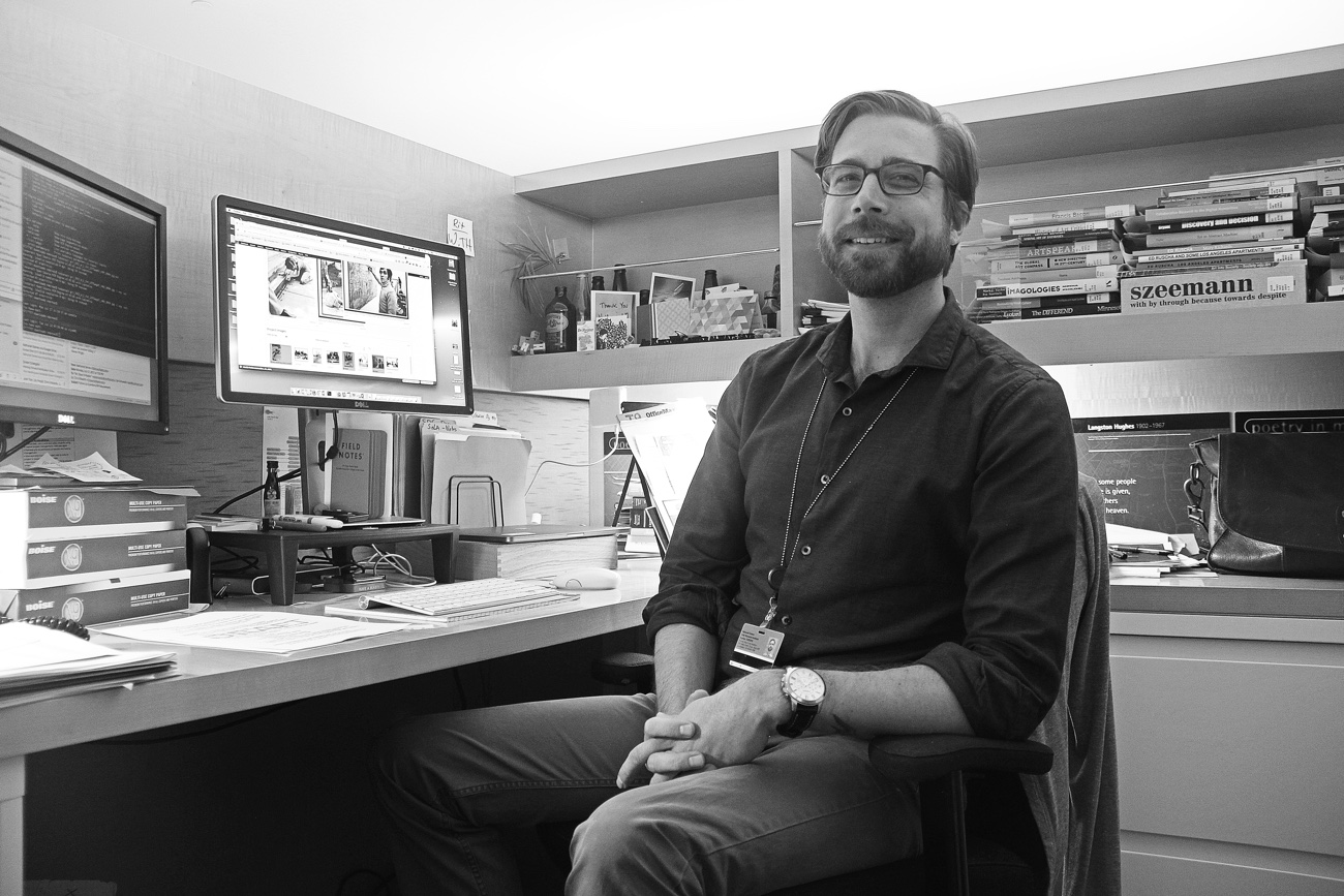 Black and white photo of a man in his late 20s with a short beard, sitting in a wood-lined cube and smiling