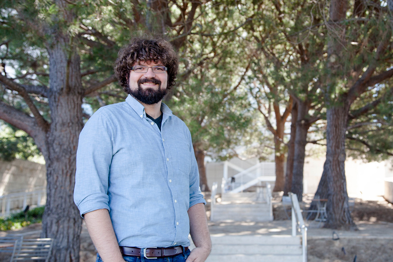 A bearded young man in a blue business shirt smiles while standing in front of several pine trees