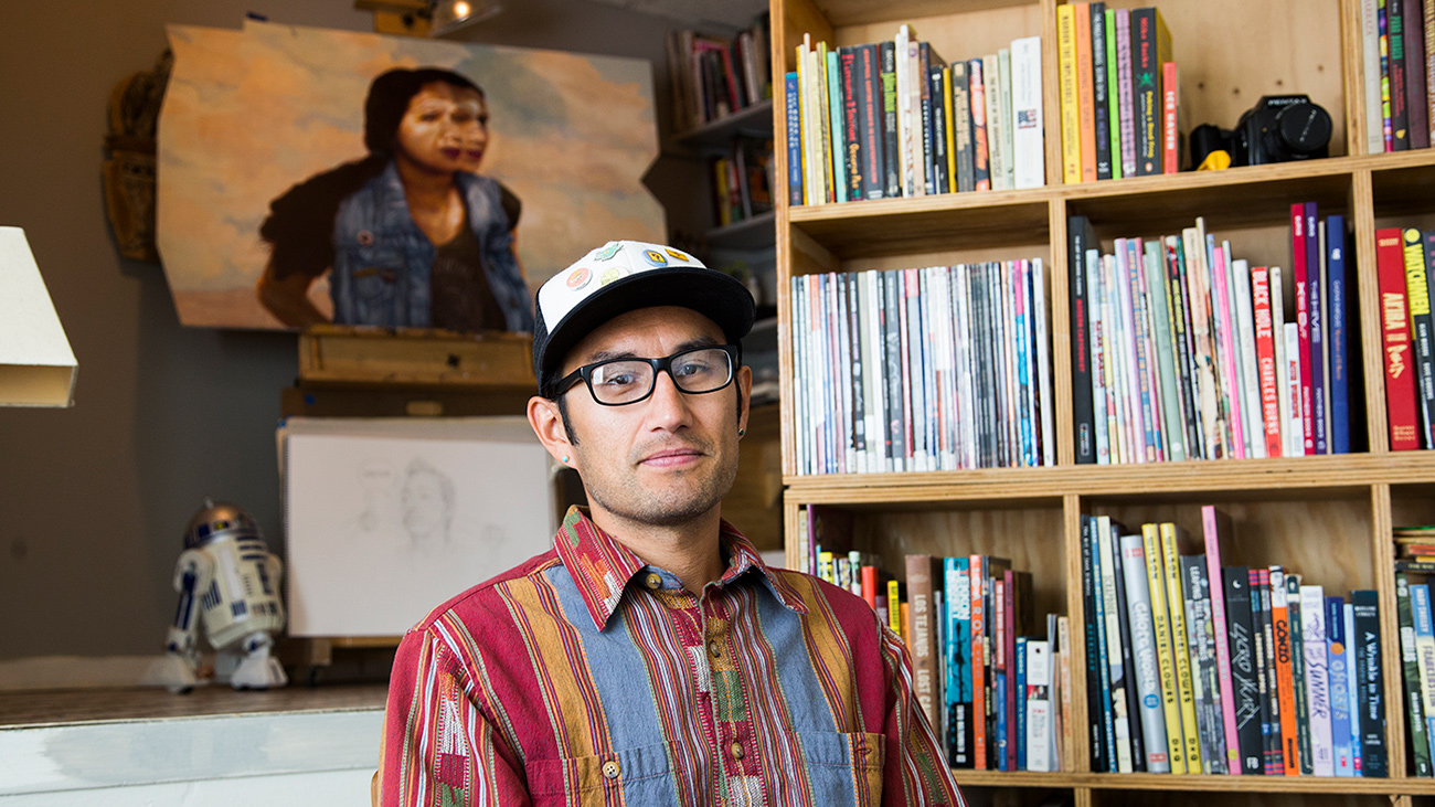 Artist and illustrator Zeke Pena sits in his studio, looking at the camera. He is wearing a colorful red, orange and blue shirt, a baseball cap and black framed glasses.