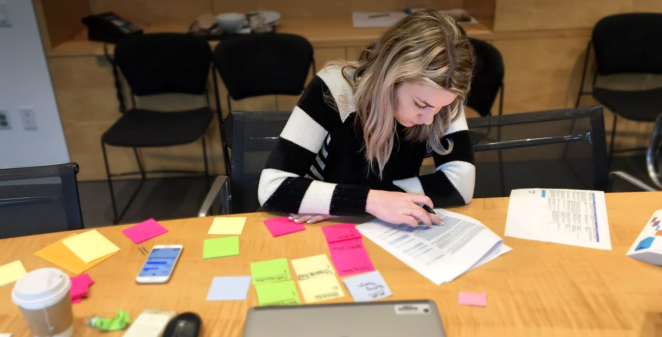A woman works at a desk in front of a closed computer, a coffee cup and papers with sticky notes on them.