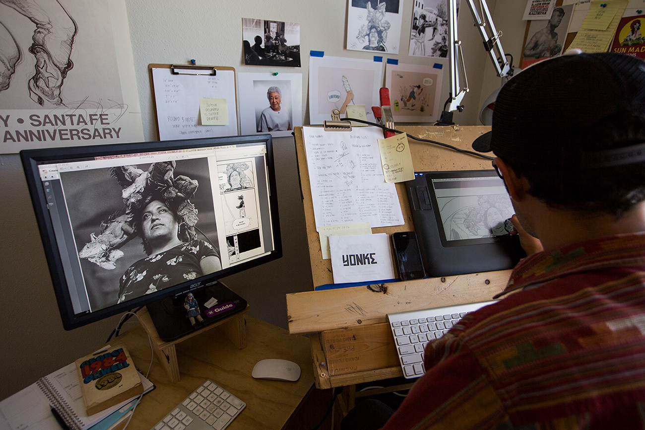 An artist has his back toward the camera drawing on a tablet from a photograph enlarged to his left on a computer monitor.