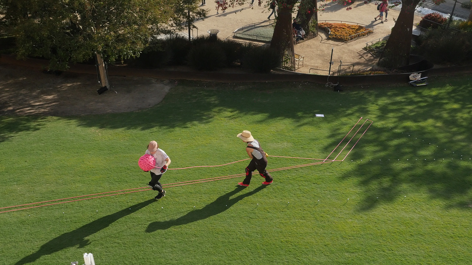Overhead view of two women on a large lawn, unspooling a large spool of bright pink rope