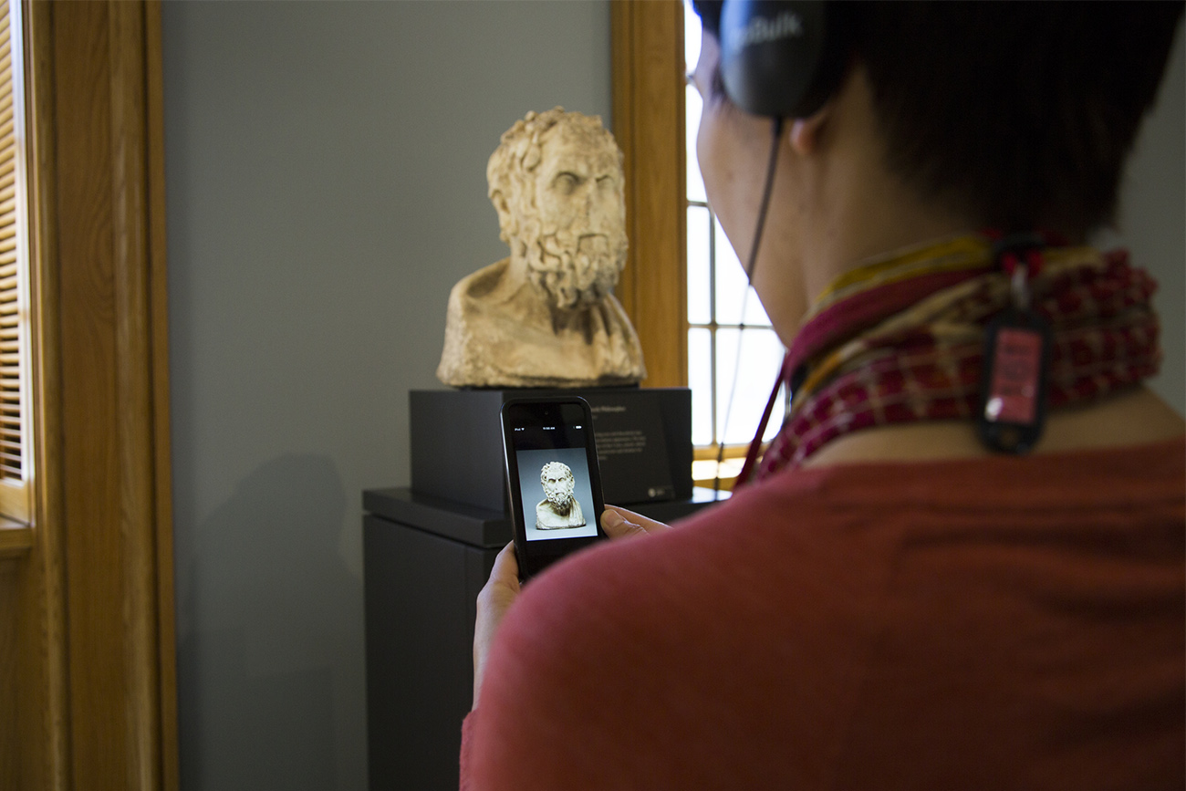 A visitor standing in front of a marble bust wears headphones and holds a GettyGuide multimedia player whose screen displays the same marble bust.