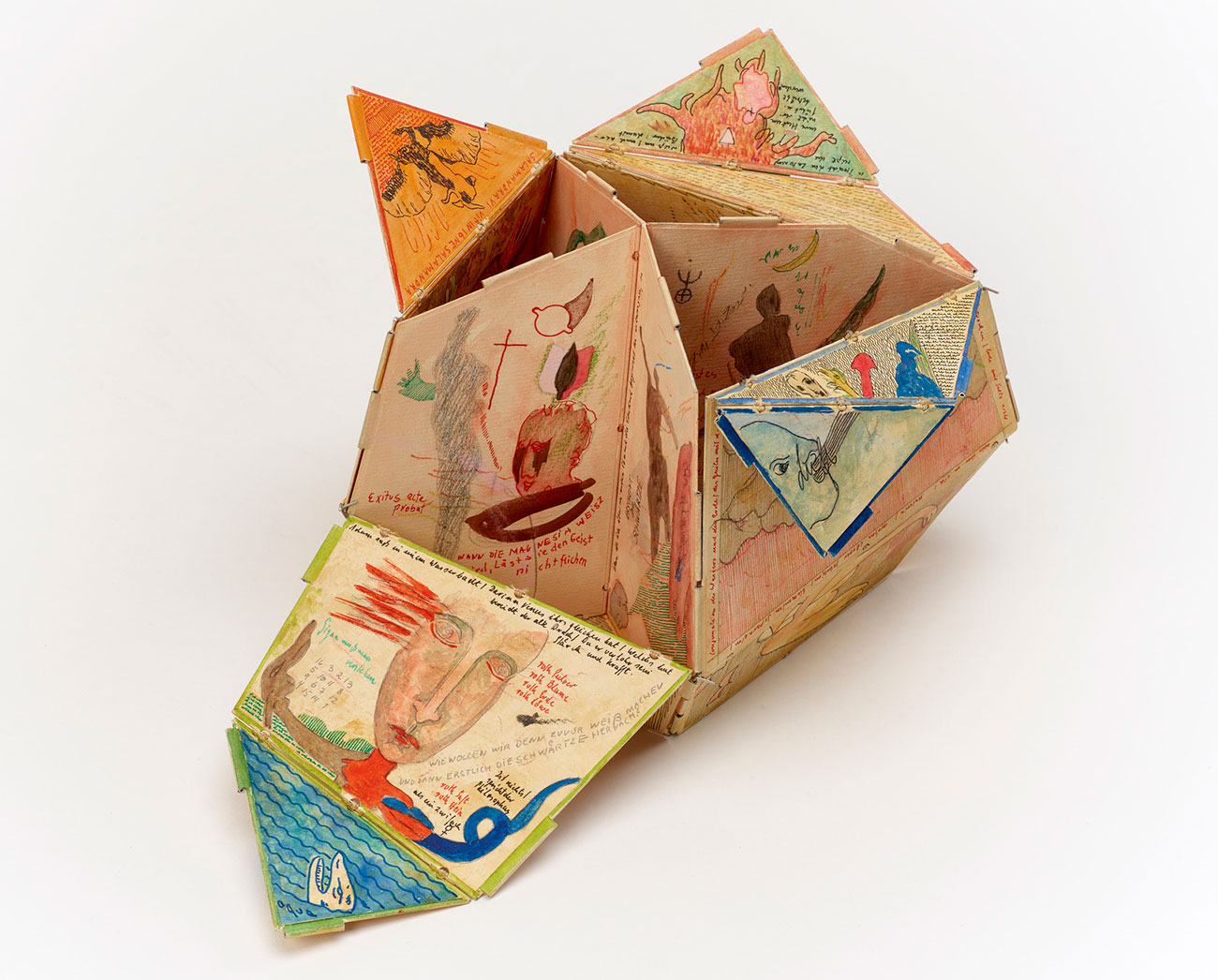 A small sculpture composed of cardboard panels decorated with hand-drawn and painted illustrations and words. Panels are irregular triangular and trapezoidal shapes and appear to open with hinges