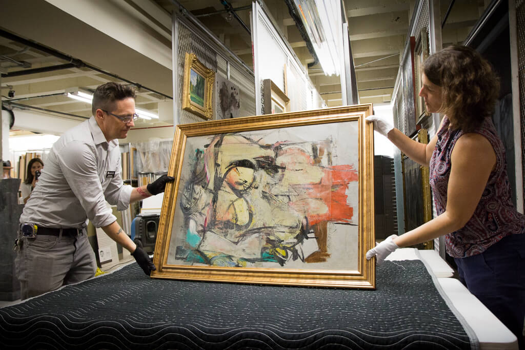 A man and a woman hold up an abstract painting featuring colors of soft gold, turquoise and coral.