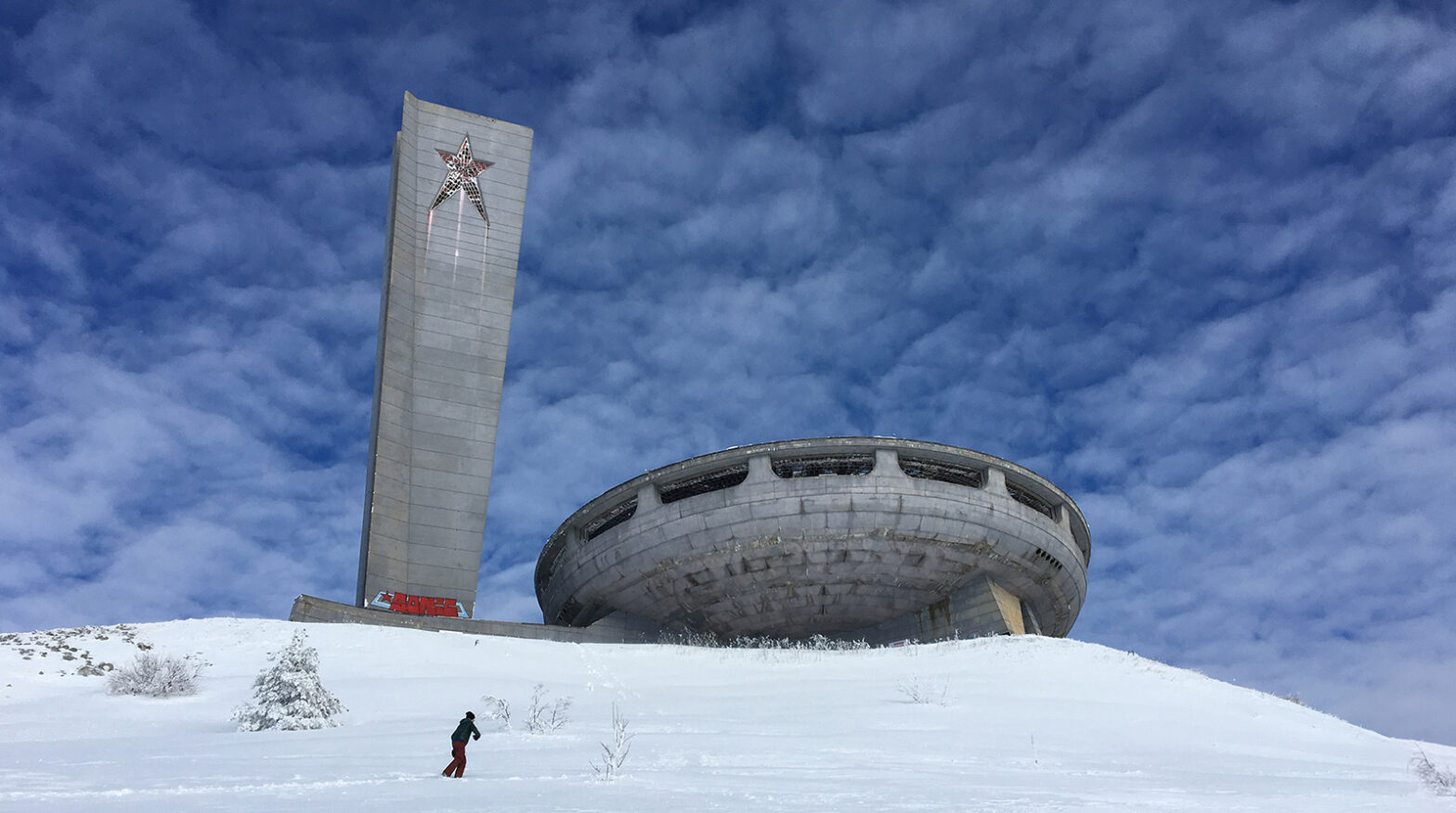 A cement spaceship-shaped building with long horizontal windows sits on a snowy hill, framed by a sky with wispy clouds. to the left is a tall cement tower with a star on it.