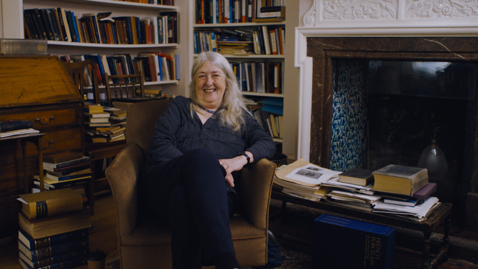 Mary Beard sits and smiles in a chair in an office full of bookshelves and books stacked everywhere.