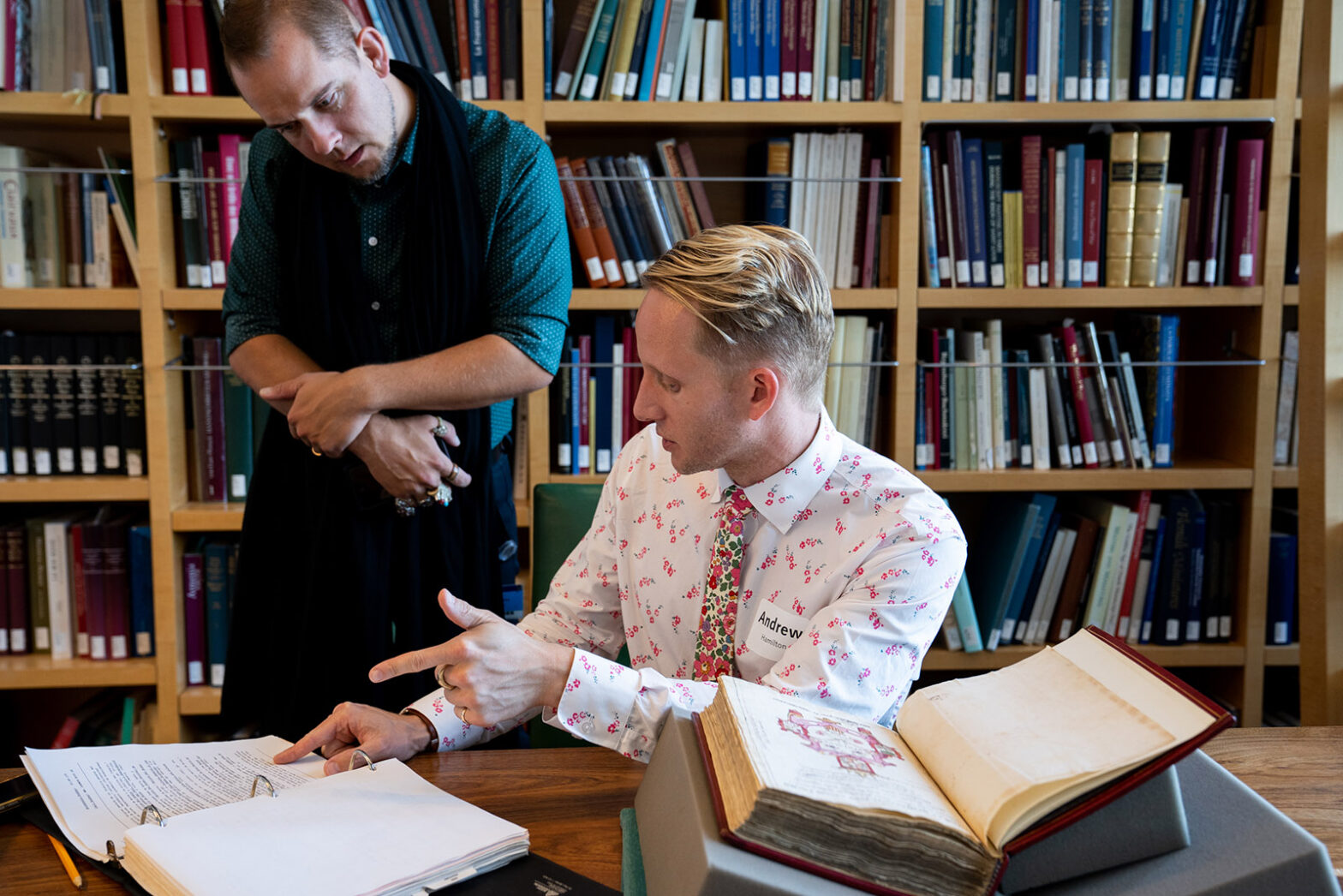 Andrew Hamilton sits at a table and points at typed pages in a binder while talking to Bryan Keene at his right.