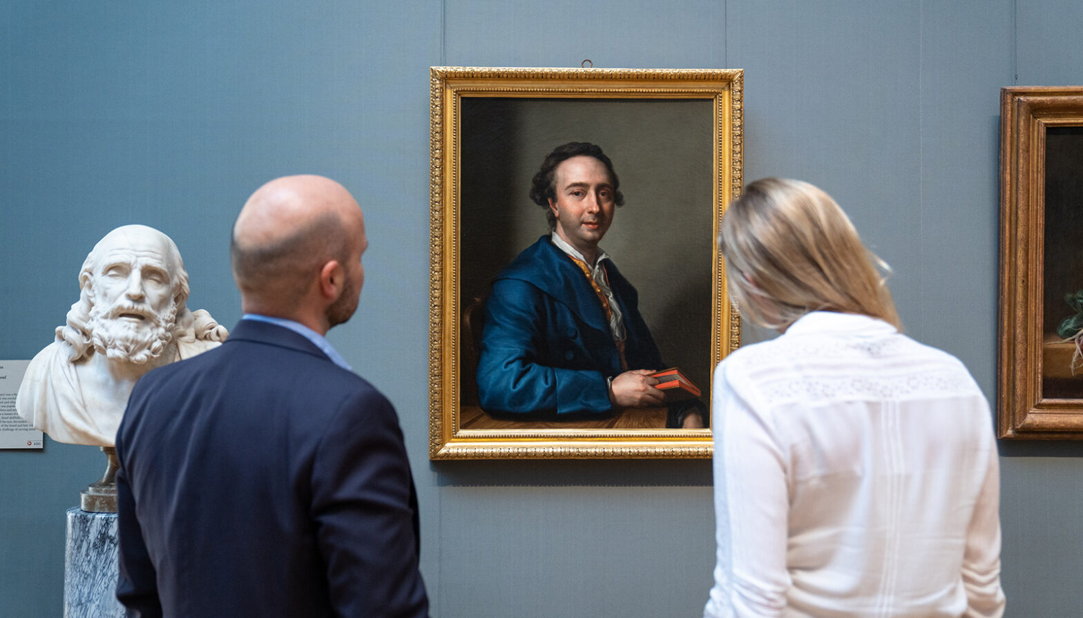 Two people looking at the new portait of a seated man in a blue coat.