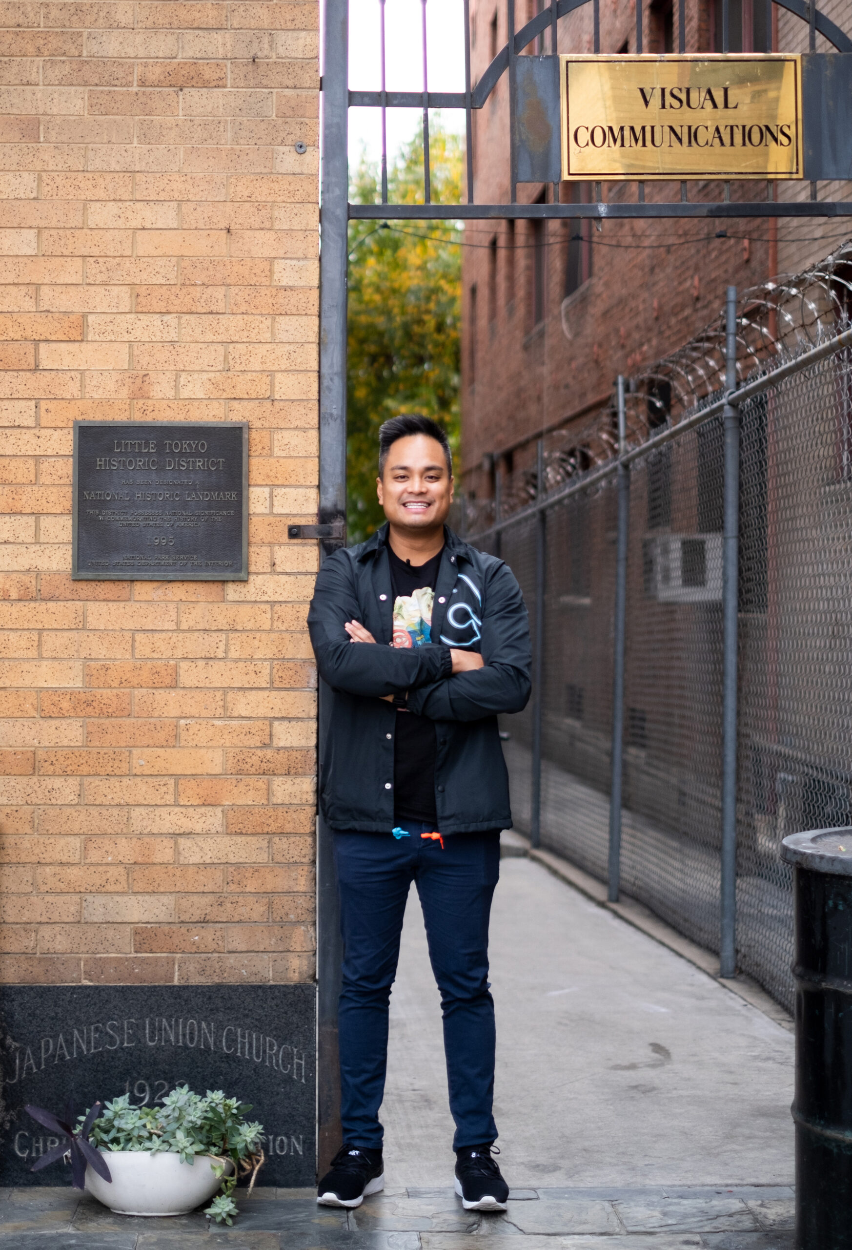 man stands next to a building with a plaque that says Little Tokyo. His arms are crossed and he smiles.