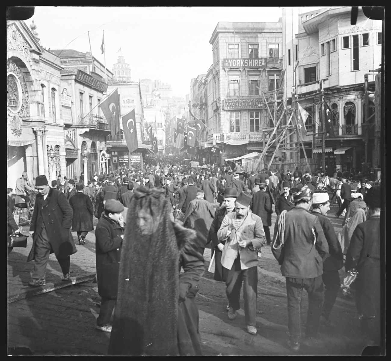 A crowd in Constantinople walking through a busy narrow street lined by buildings that are 2 and 3 stories tall. A woman in the foreground has her face covered by a sheer black veil.