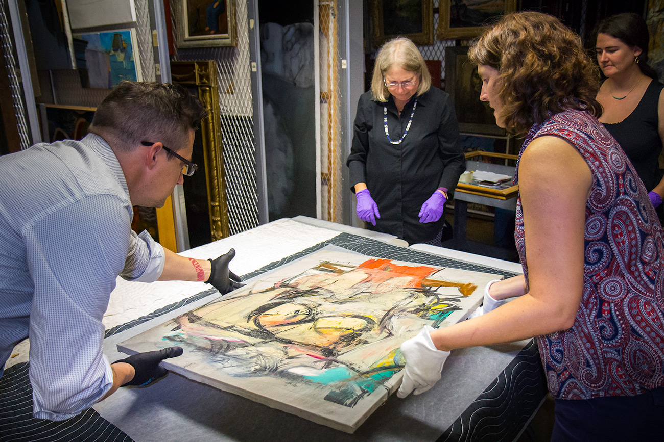 Three conservators examine an abstract painting featuring colors of soft gold, turquoise, and coral. Willem de Kooning, Woman-Ochre, 1955. Oil on canvas, 30 x 40 in. Copyright 2019 The Willem de Kooning Foundation / Artists Rights Society (ARS), New York