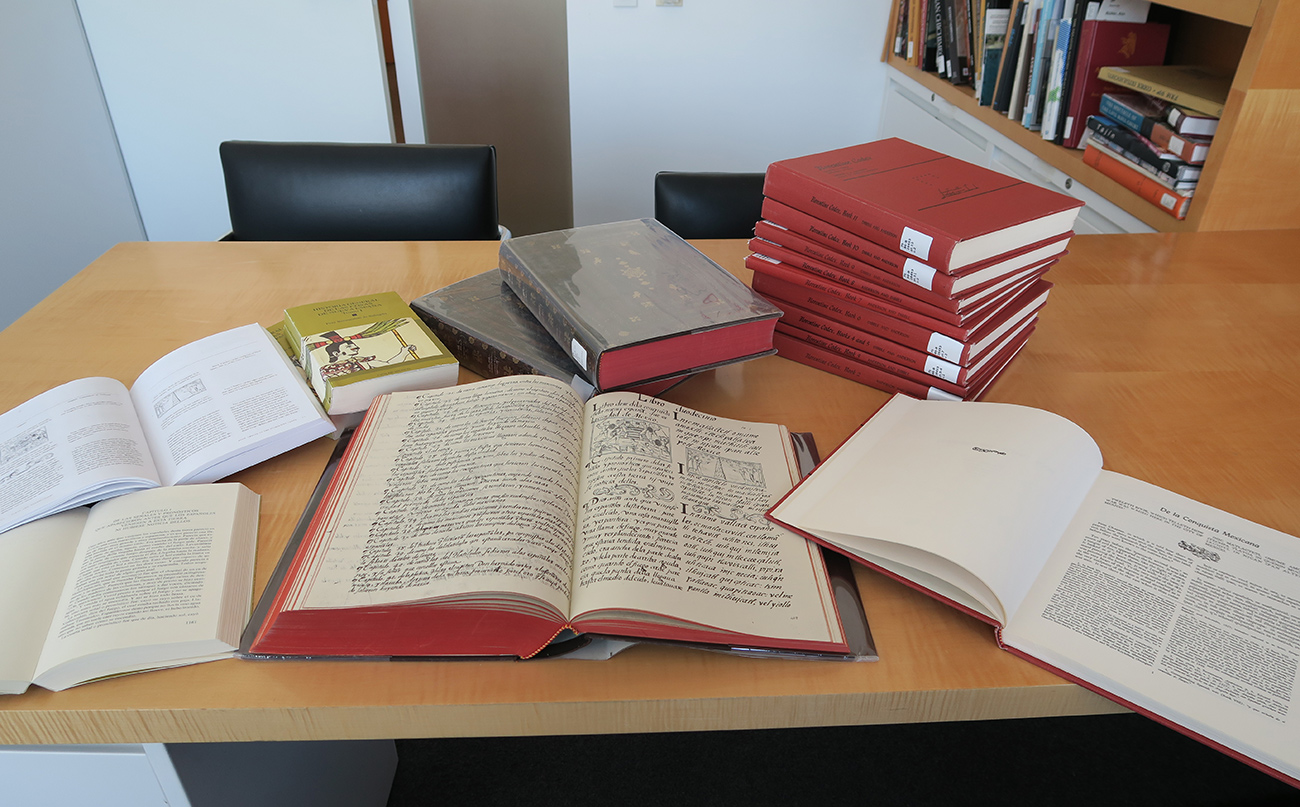 Books, some opened, some stacked, on a light-wood table.