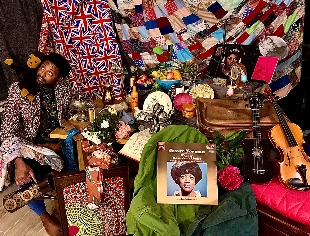 Brathwaite sits with a stuffed Curious George on his shoulder, in a room filled with a lot of stuff, including musical instruments, albums, and fruit. Quilts and blankets hang in the back.