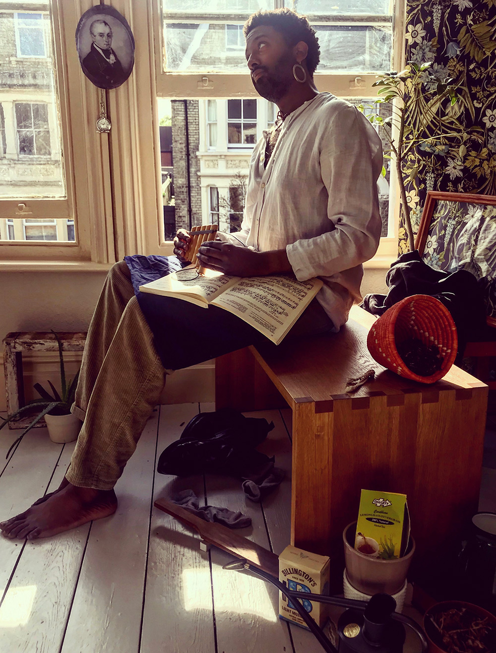 Brathwaite sits by two windows, one opened. He's wearing a white shirt and holding a book of sheet music and a musical pipe.