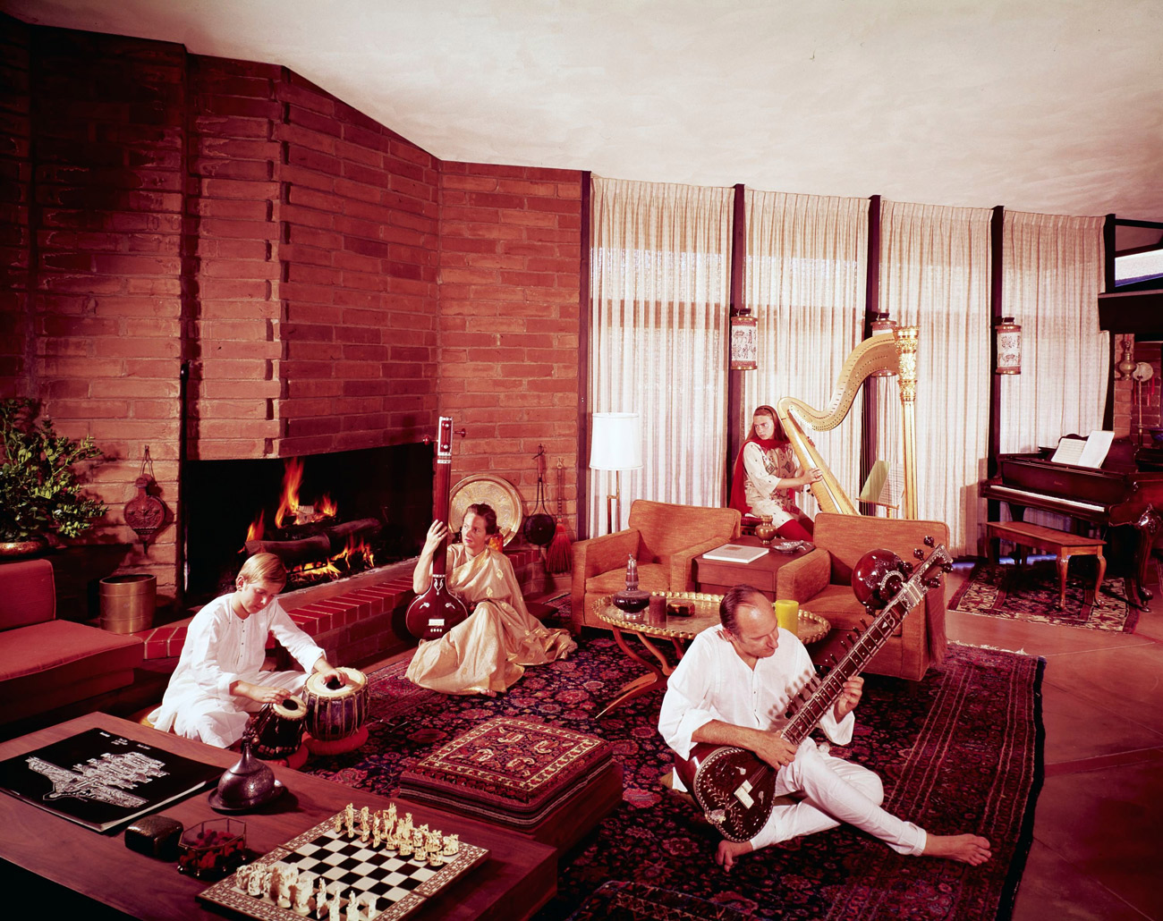 Four musicians with classical instruments, including a large harp, are seated in an expansive living room decorated in shades of red