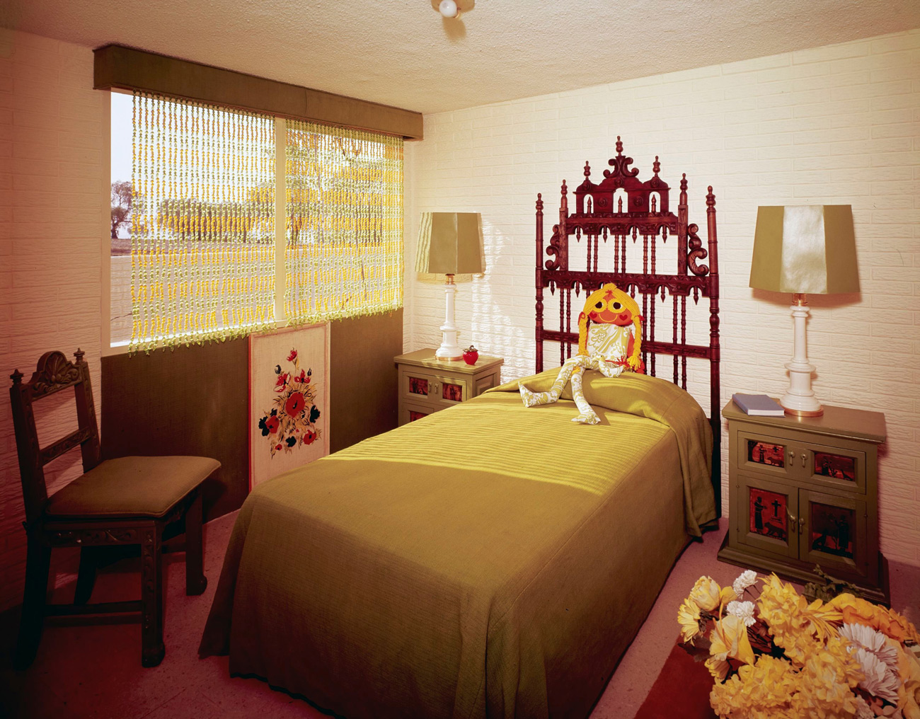 Small white bedroom with olive green bedspread and accents. A doll sits on the pillow.