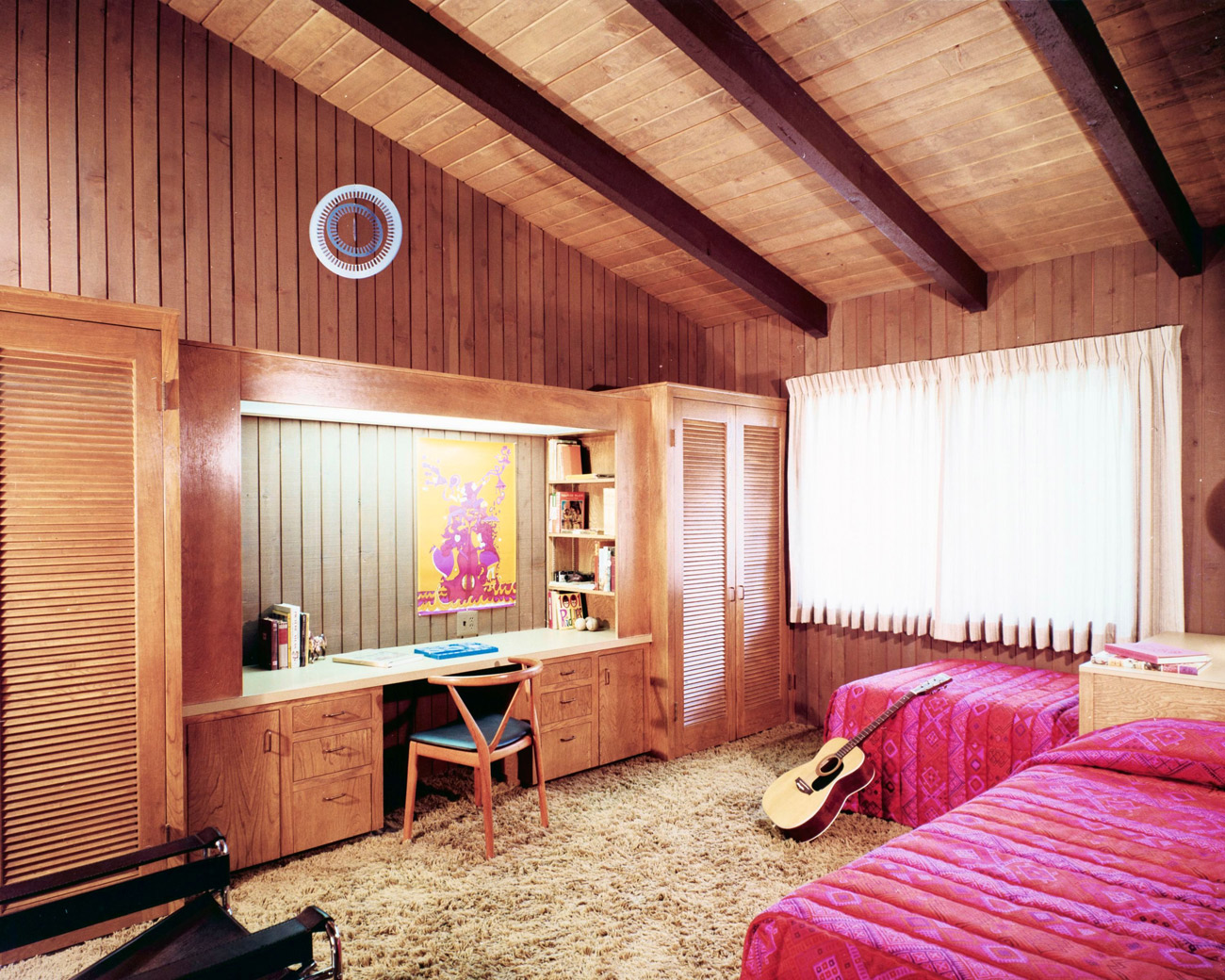 Wood paneled bedroom with slanting wood ceiling, two twin beds with pink linens, a desk and an acoustic guitar