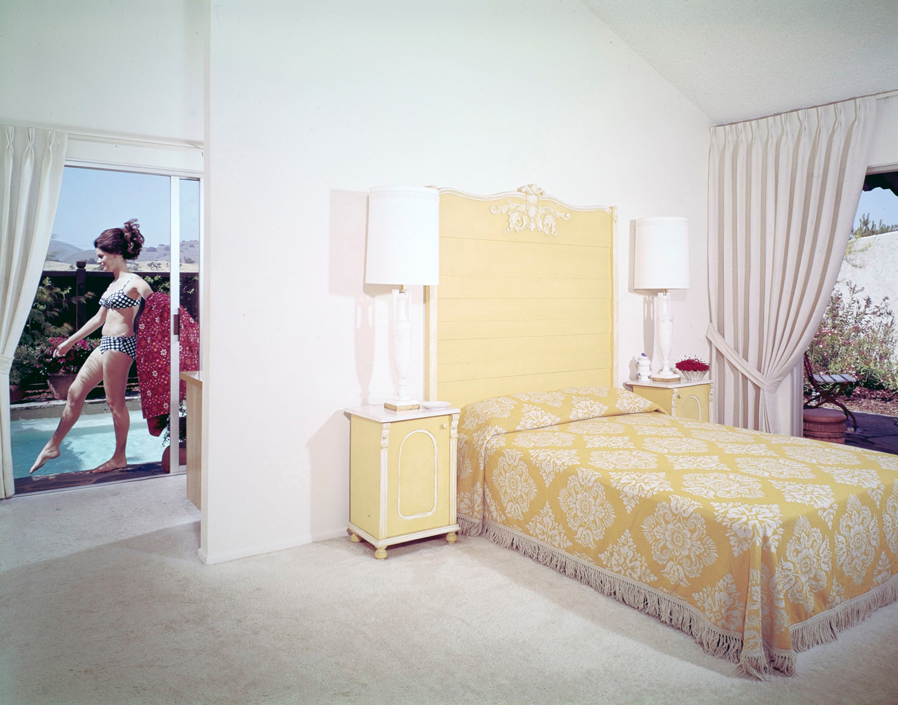 White bedroom with yellow accents. A woman is seen through a window by the pool.