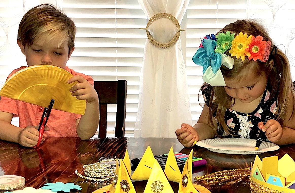 Two children sit at a table making crowns with paper plates