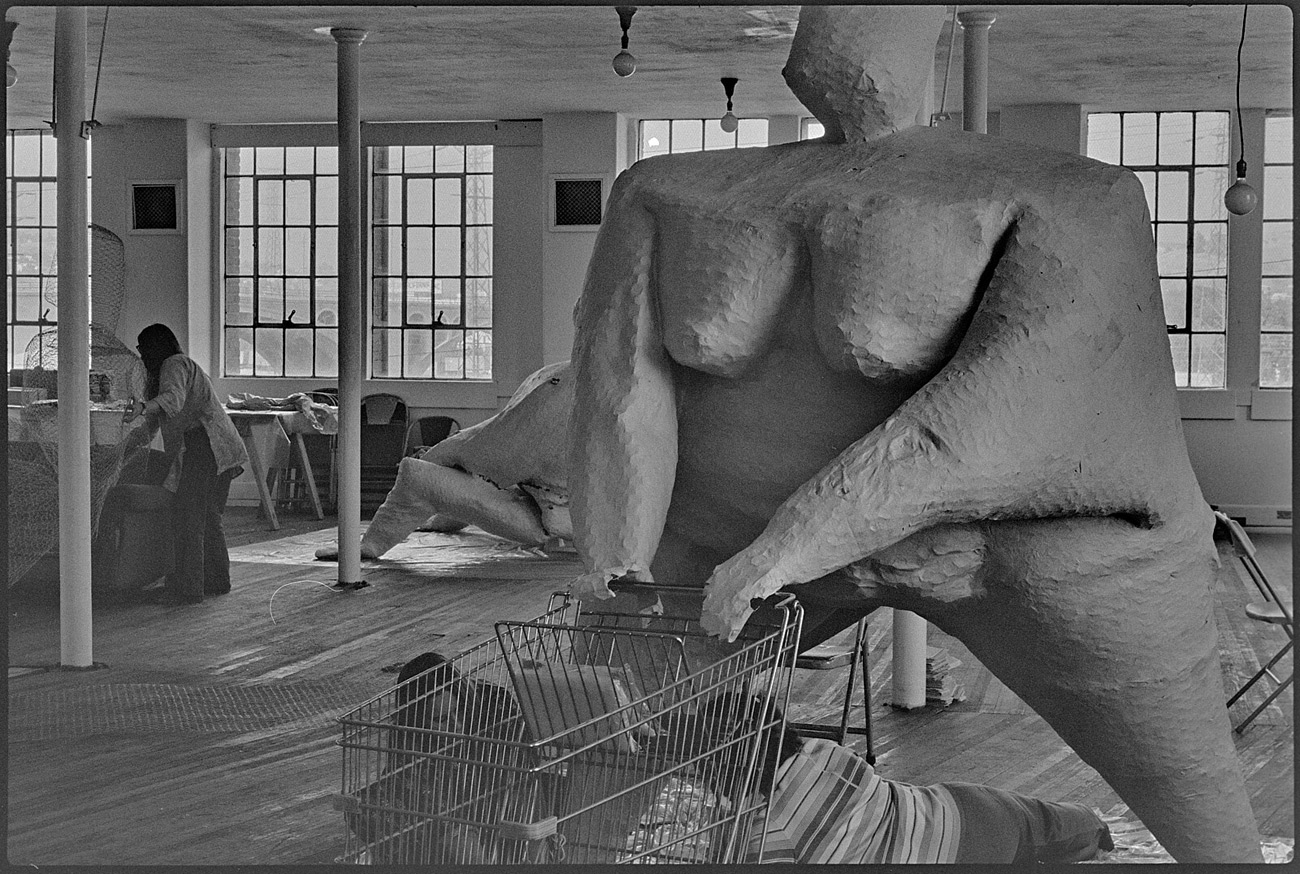 Large sculpture of nude woman with hands on a shopping cart. Two people lie on floor working on a leg.