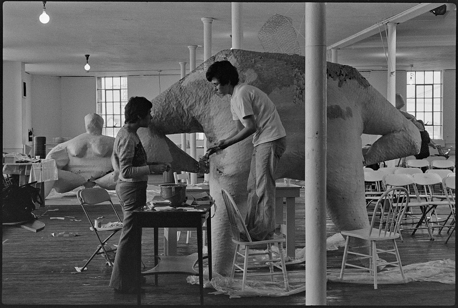 Two people work on large sculpture while another sculpture sits in the background