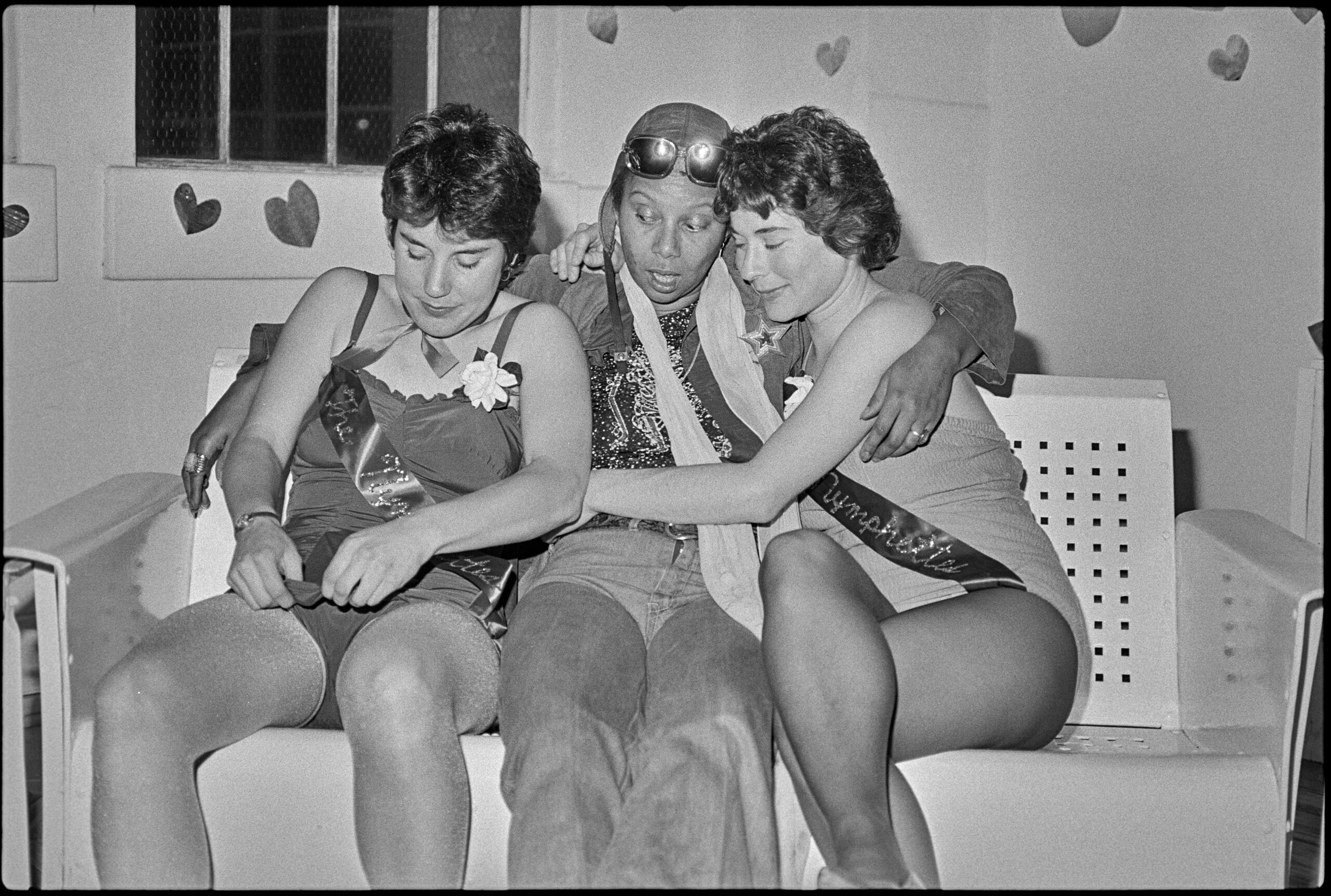 Three women sit with their bodies entwined on a couch. There are small paper hearts on the wall behind them.