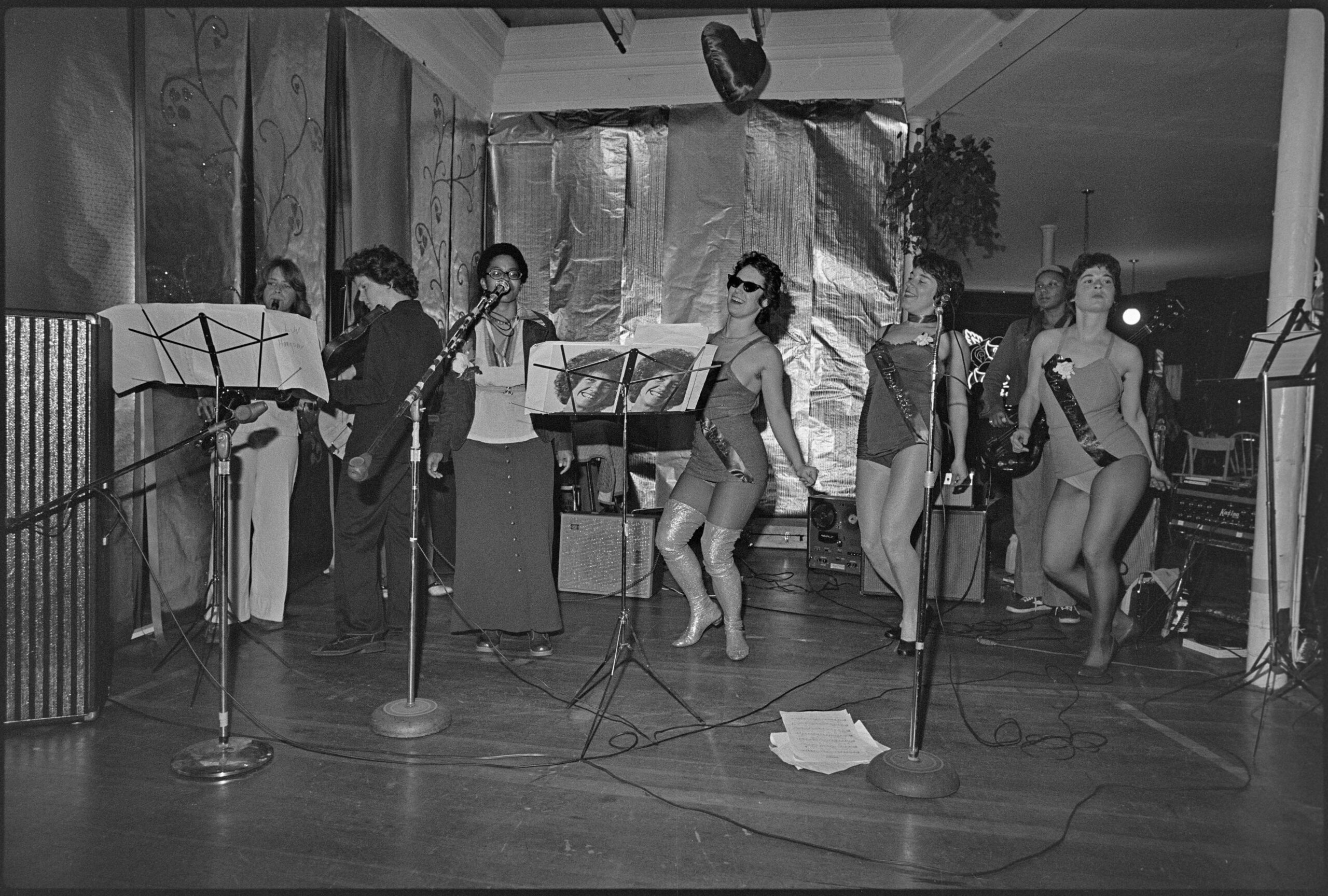 Women in leotards and sashes dance across a stage towards microphones. A band plays behind them.