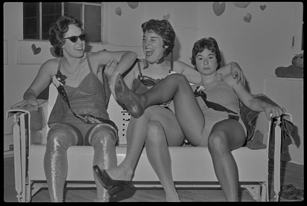 Three women sit on a couch smiling and laughing. The woman on the right reaches her foot to the left, and it is held by the woman on the left in a playful gesture.