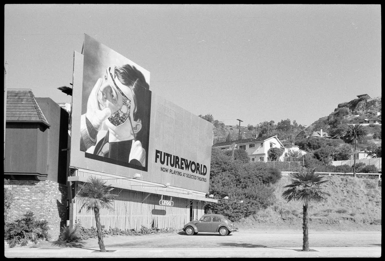 A large billboard that says Futureworld, Now playing at selected theaters, with a picture of a robot with a hand removing its face, on the side of a road surrounded by trees and houses
