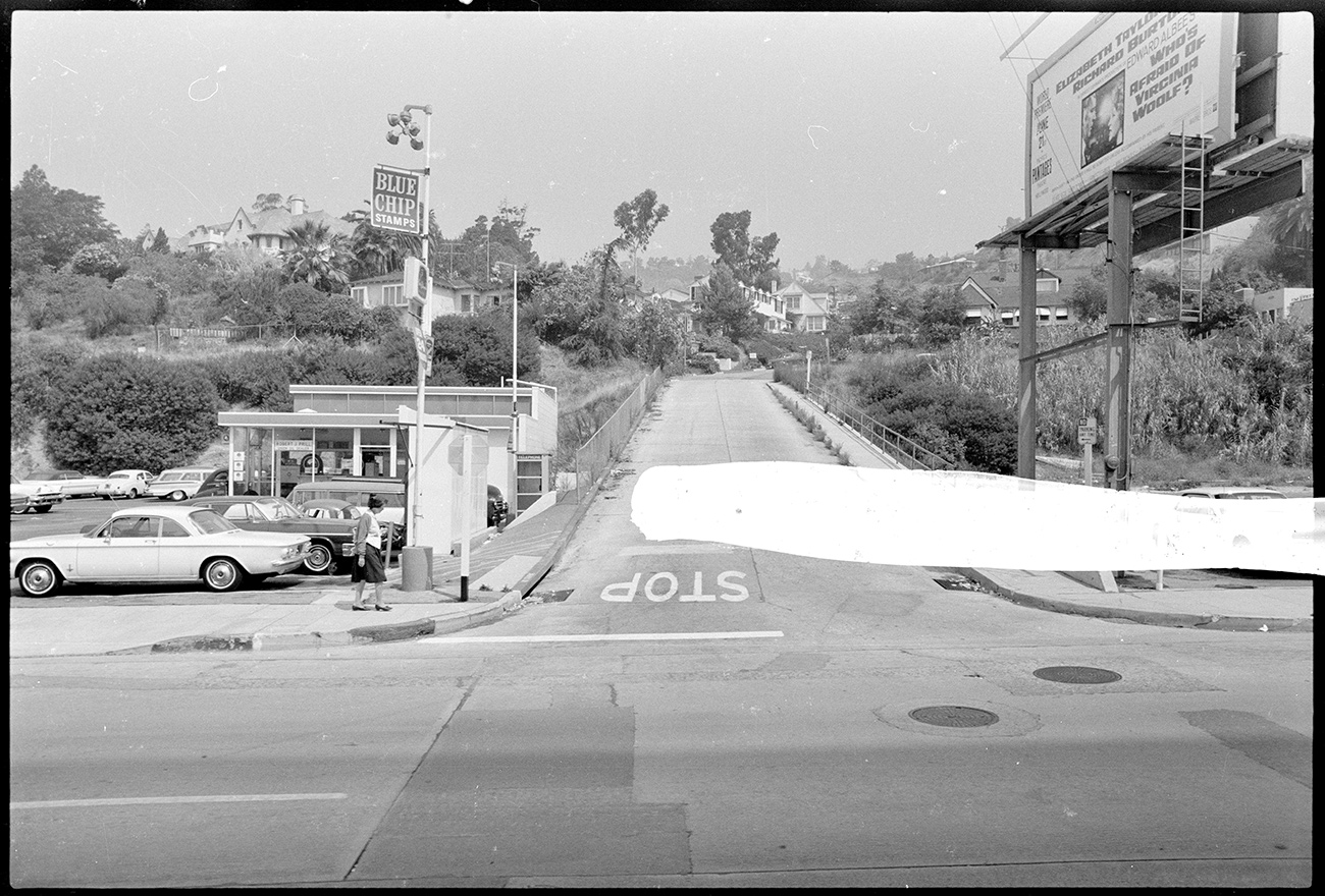 An intersection between two streets, with trees on both sides and a large white paint streak down the center of the image