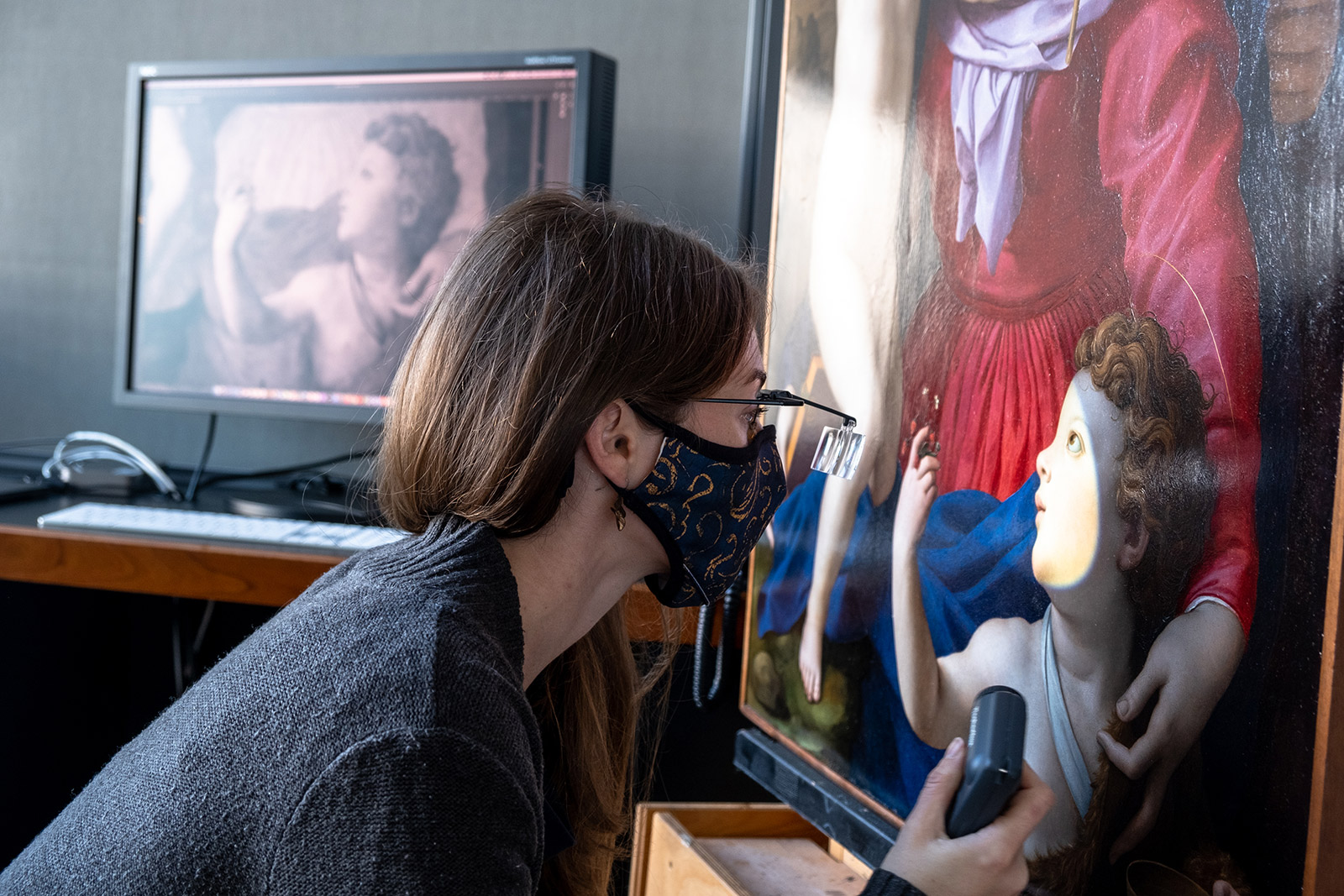 Woman wearing a face mask and glasses with magnification looks closely at a painting of a woman wearing red dress and child with curly hair