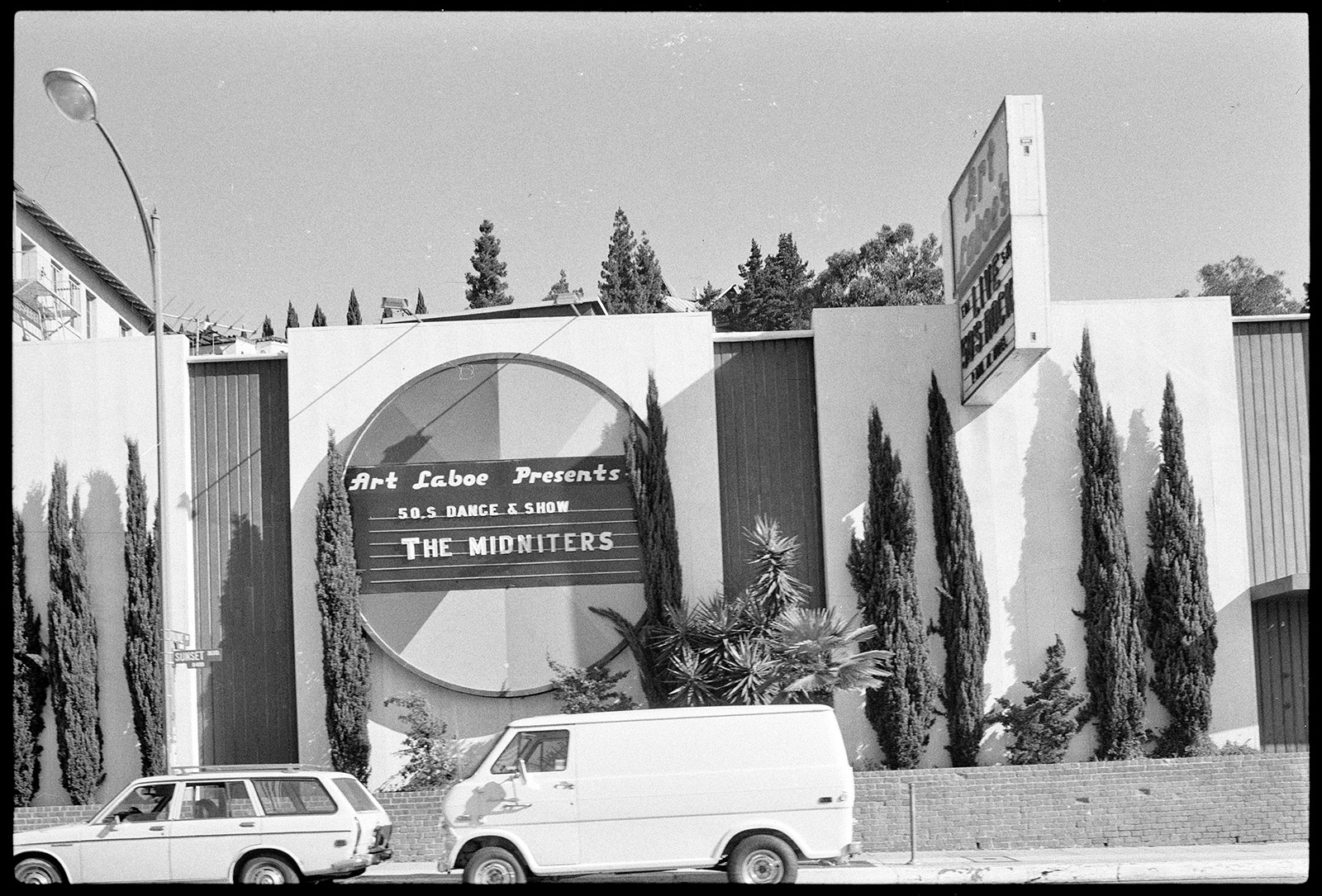 Exterior of windowless building with circle shaped sign that says Art Laboe Presents 50s Dance & Show The Midniters; there is a row of skinny trees in front of the building and a marquee that says Art Laboe’s