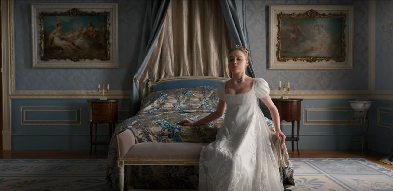 A woman wearing a flowing white dress sits on a bench in front of an ornate bed, on the wall behind the bed are two paintings that depict Venus and Cupid
