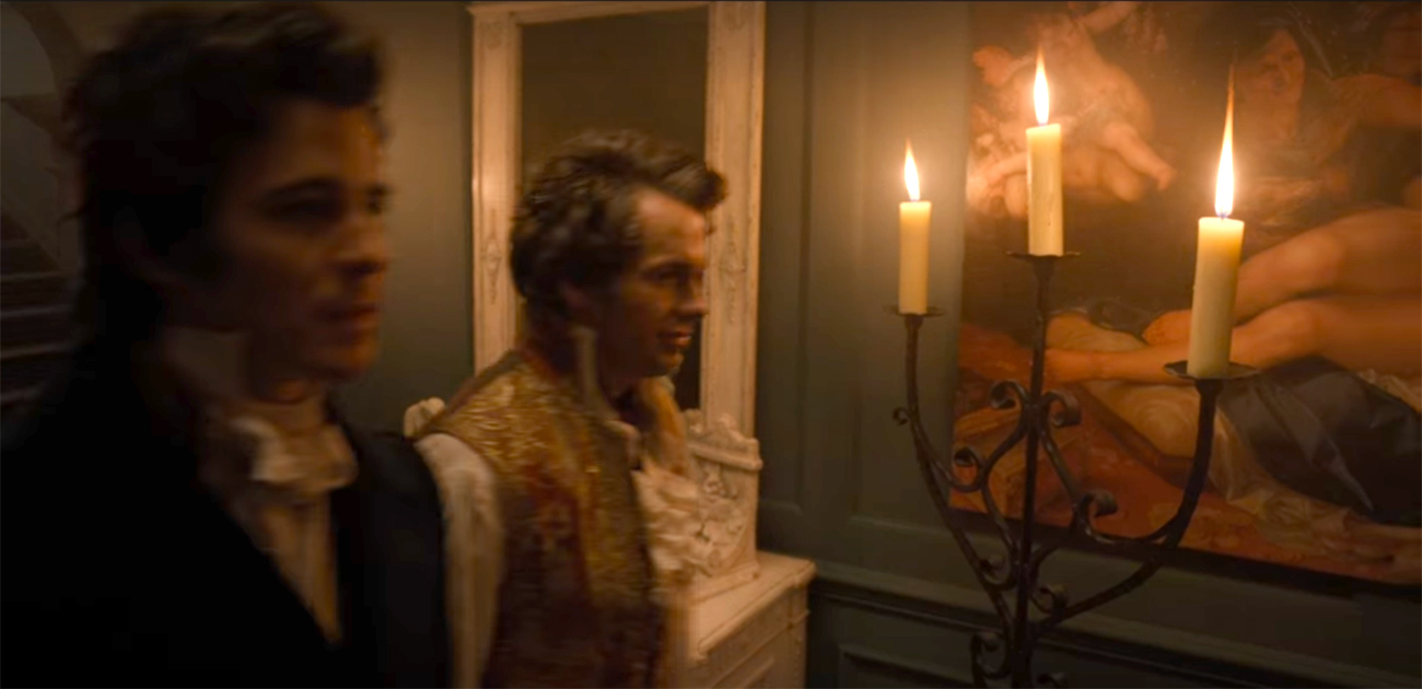 Still image from Bridgerton that shows two men standing in front of a wall, on which a mirror and a painting is hanging; you can only see half of the painting depicting a woman’s legs