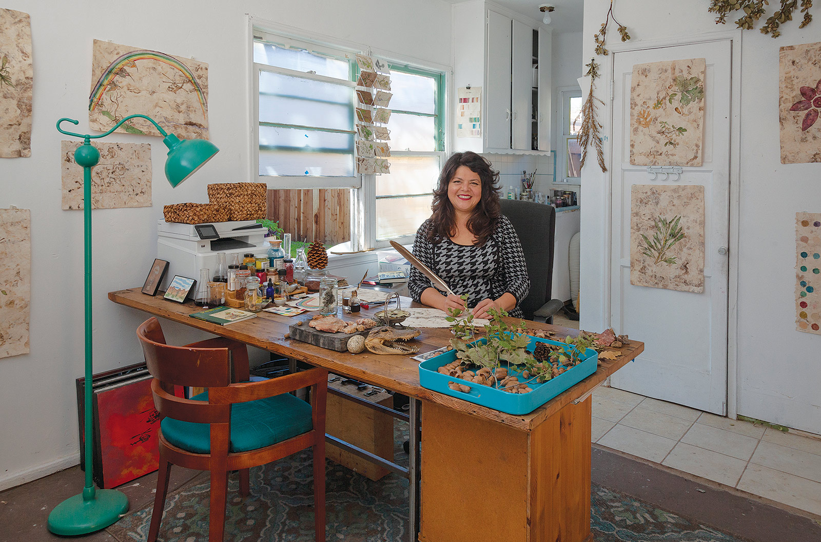 Woman sitting behind desk that has art supplies, plants, leaves, and rocks on it; there are drawings of plants on the walls around her