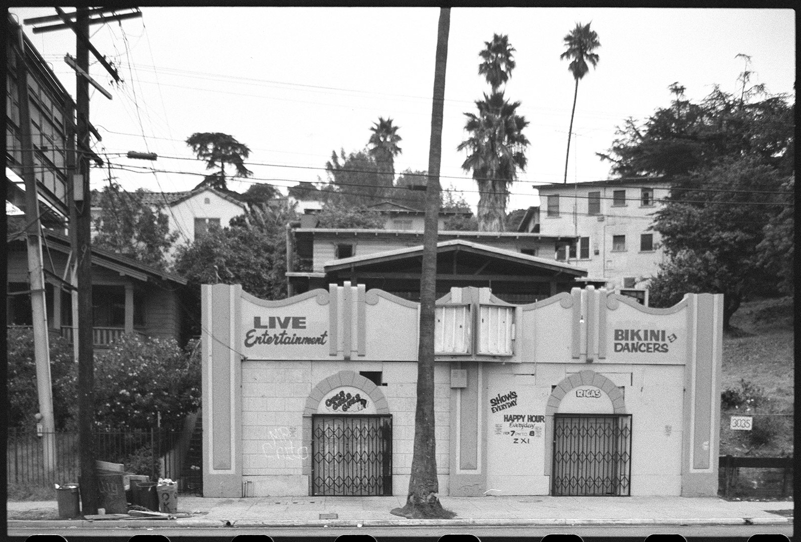 One-story building with signs that say Live Entertainment and Bikini Dancers, with gates over two doors at the front and trees and apartment buildings behind the building