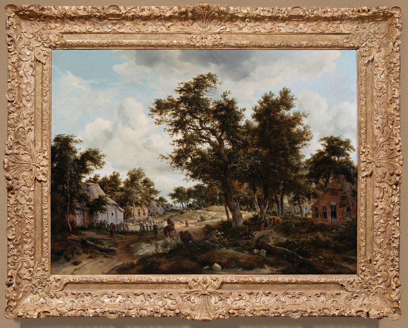 Painting of country road featuring cottages and leafy trees, framed in pale brown frame adorned with curled, scalloped detailing