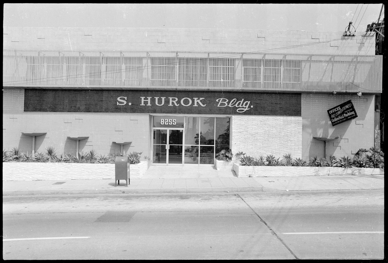 Street view of a building that features lettering that says S. Hurok Building, with a row of windows at the top of the building and a panel of floor to ceiling windows next to the glass door