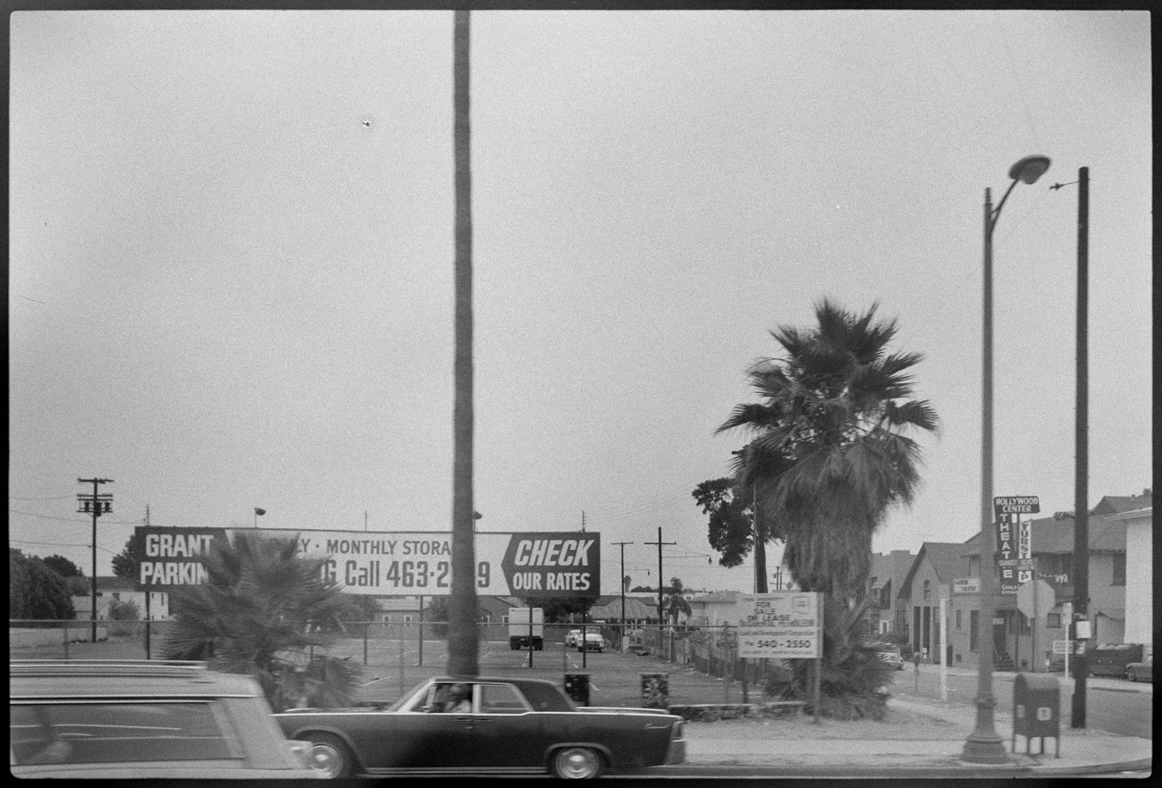 Parking lot surrounded by chain link fence with sign that says Grant Parking, Monthly Storage, Check our rates. There are palm trees next to the lot and apartment buildings on the other side of the street