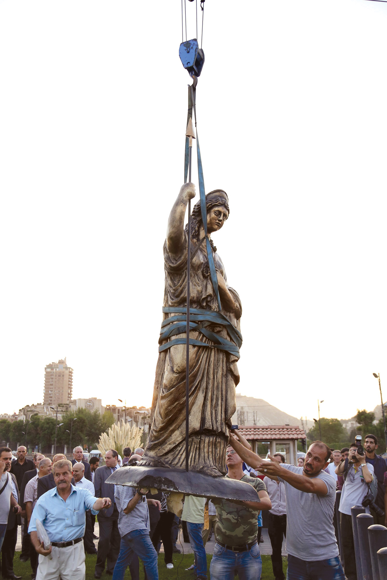 Group of men lifting a statue of a woman wearing a crown and draped in robes, using cables and a crane