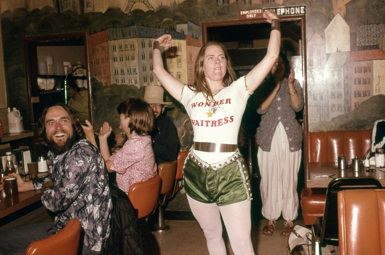 woman stands in a diner with her arms up and a t-shirt that says wonder waitress, while onlookers smile