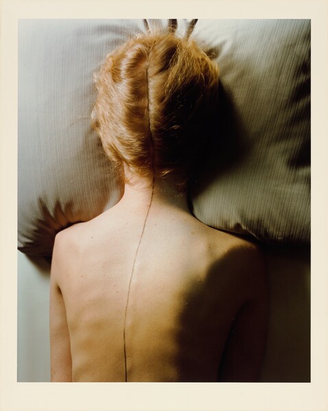 Woman lying facedown on a pillow, with her blonde hair split into two sections and folded close around her head and a pencil line drawn down her spine