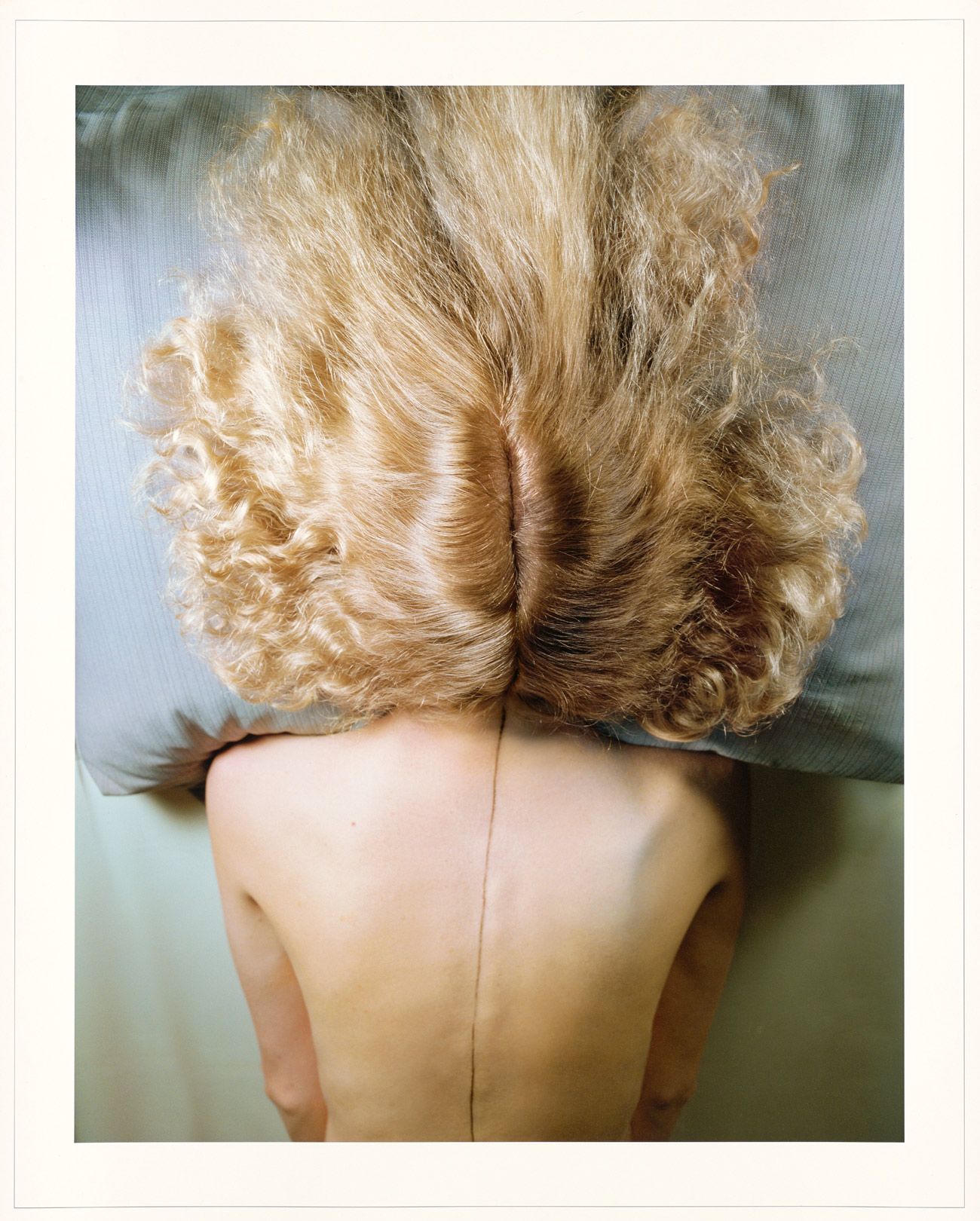 Woman lying facedown on a pillow, with her blonde hair split into two sections on either side of her head and flowing onto the pillow, and a pencil line drawn down her spine
