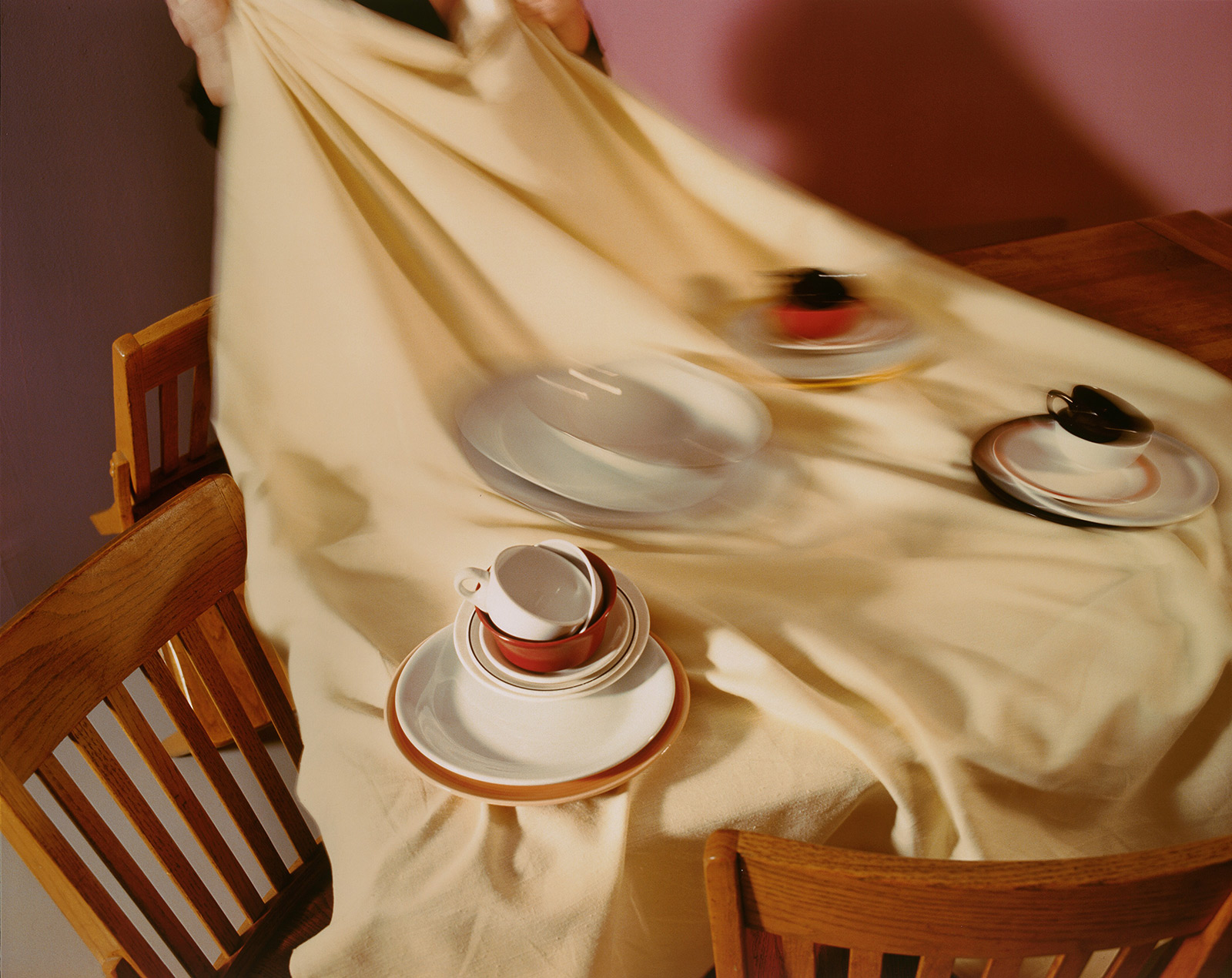 A tablecloth is pulled off a table surrounded by wooden chairs; the tablecloth and stacks of plates, saucers, and teacups on top are blurry
