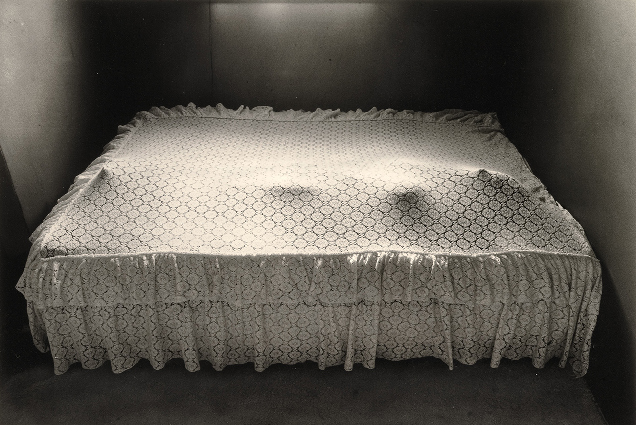 A person lies under a white patterned bedsheet creating impressions with their arms and legs