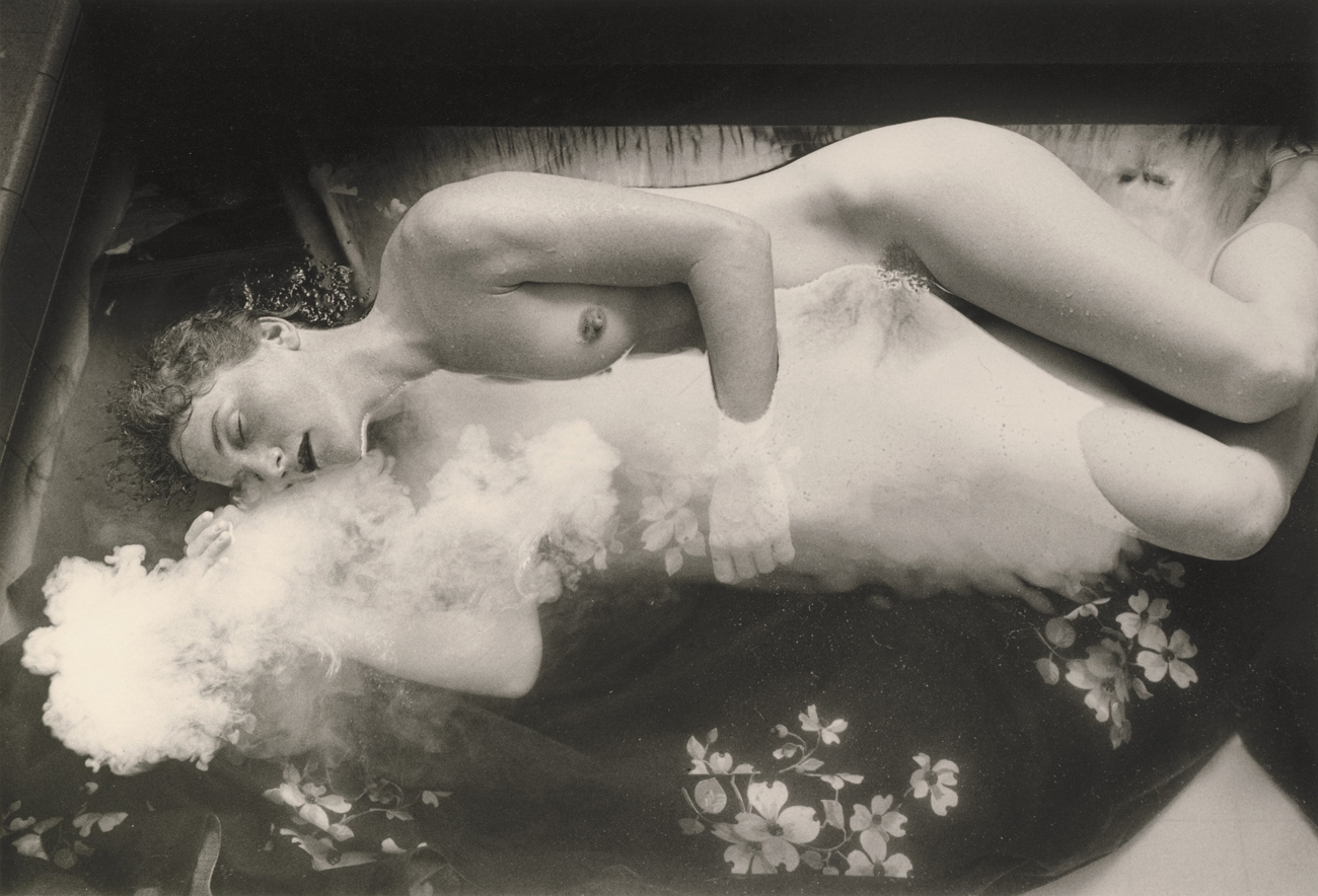 Nude woman lying on her side partially submerged in water, on a floral fabric, with her arms around white clouds
